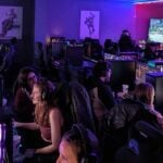 Busy esports center interior with rows of gamers playing on PCs under dim purple ambient lighting.