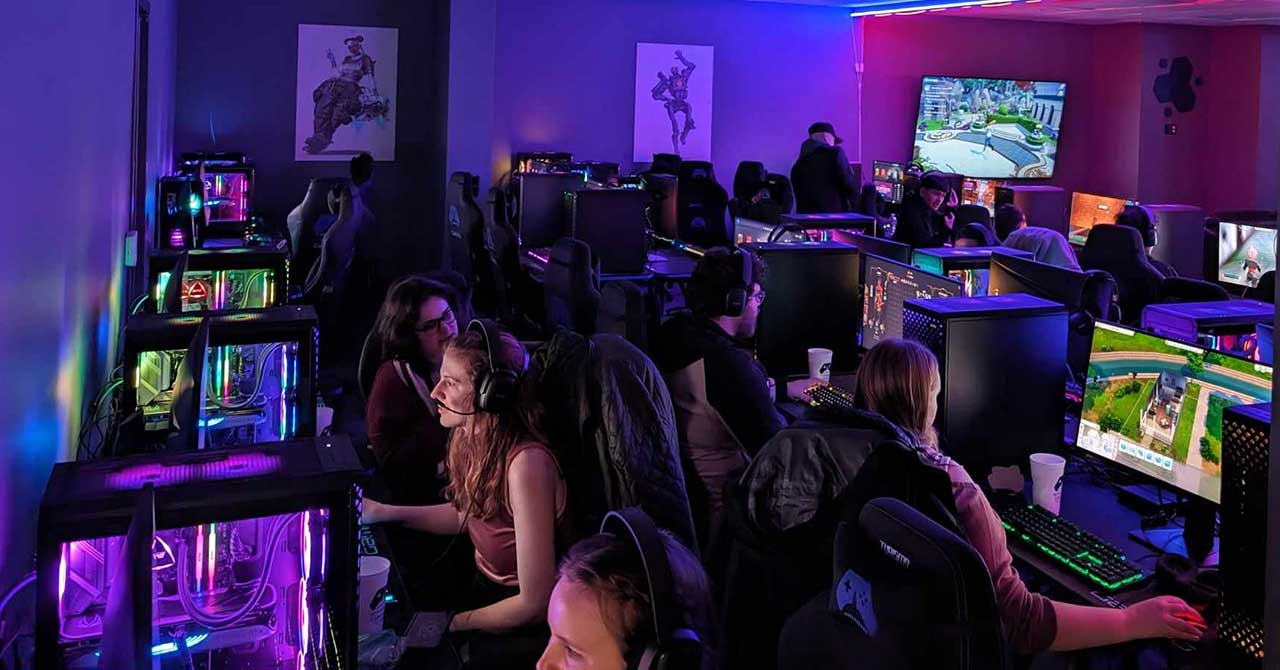 Busy esports center interior with rows of gamers playing on PCs under dim purple ambient lighting.